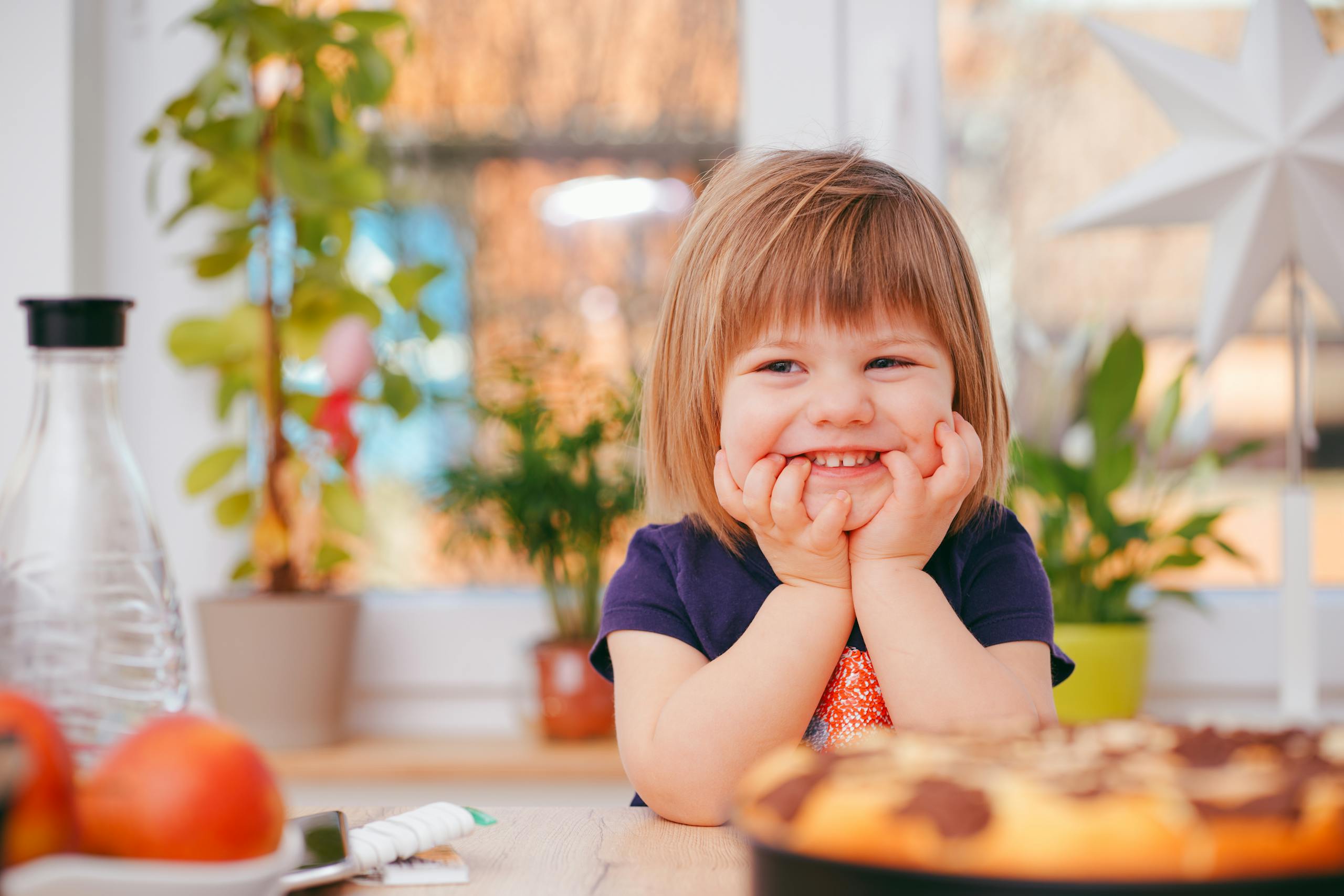 A happy young girl smiling indoors surrounded by plants and a pastry, with natural light streaming in.